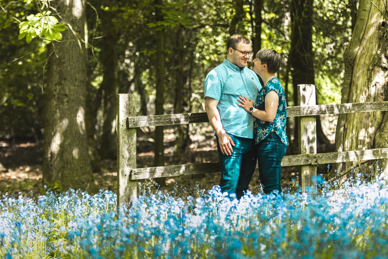 Two people in matching teal shirts stand together by a rustic fence surrounded by blooming blue wildflowers in a wooded setting.