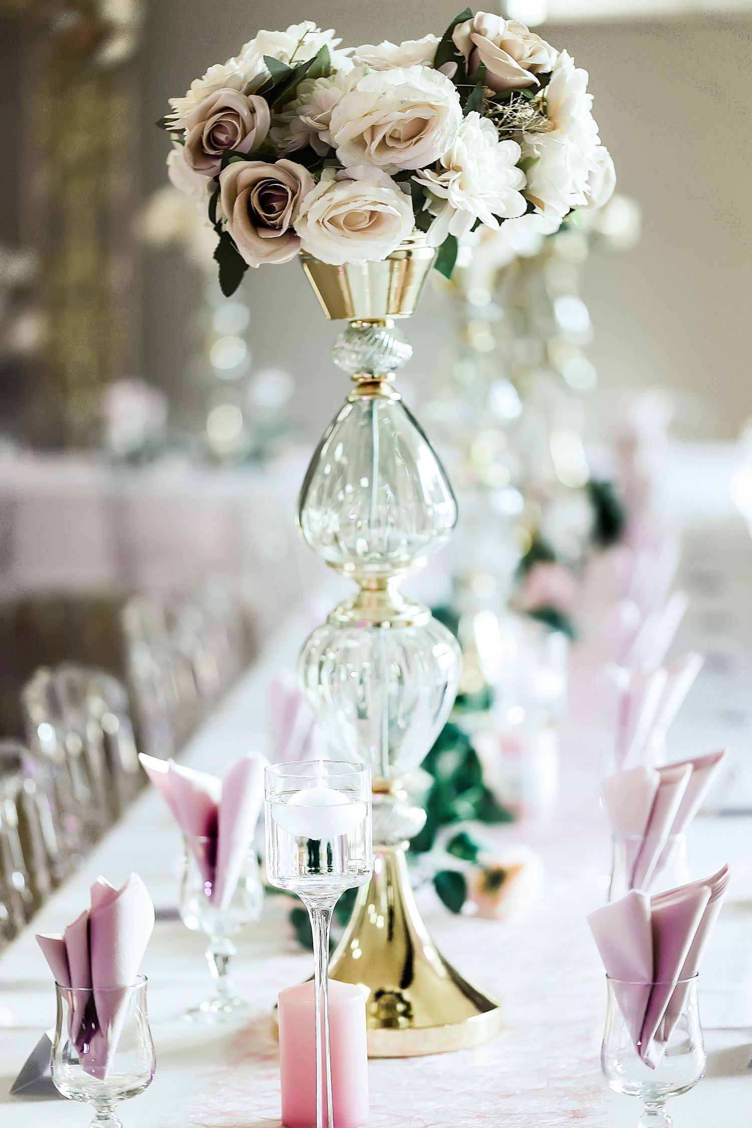 Elegant wedding centerpiece with blush and white roses in a tall gold and glass stand atop a white tablecloth with pink napkins.