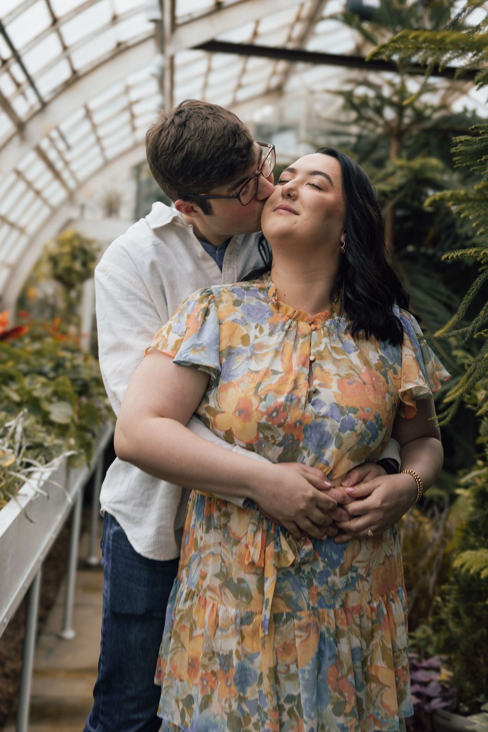 A romantic couple embraces in a sunny greenhouse conservatory surrounded by lush greenery and natural light.