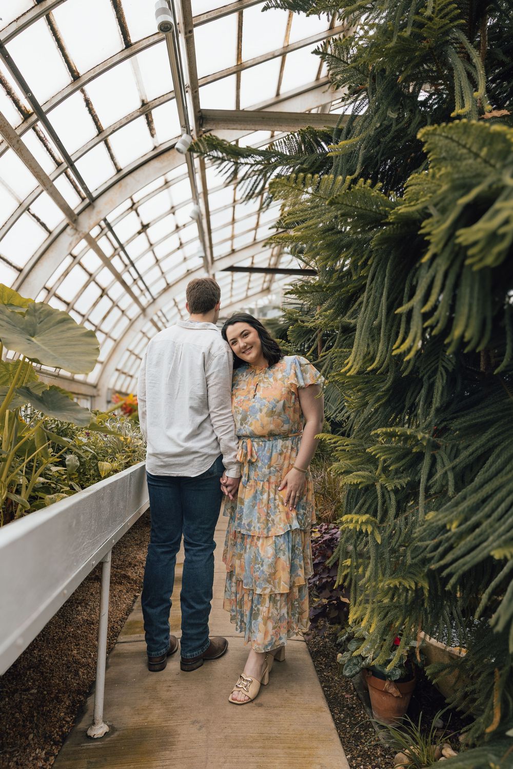 A couple walking through a greenhouse pathway lined with tropical plants and a curved glass ceiling overhead.