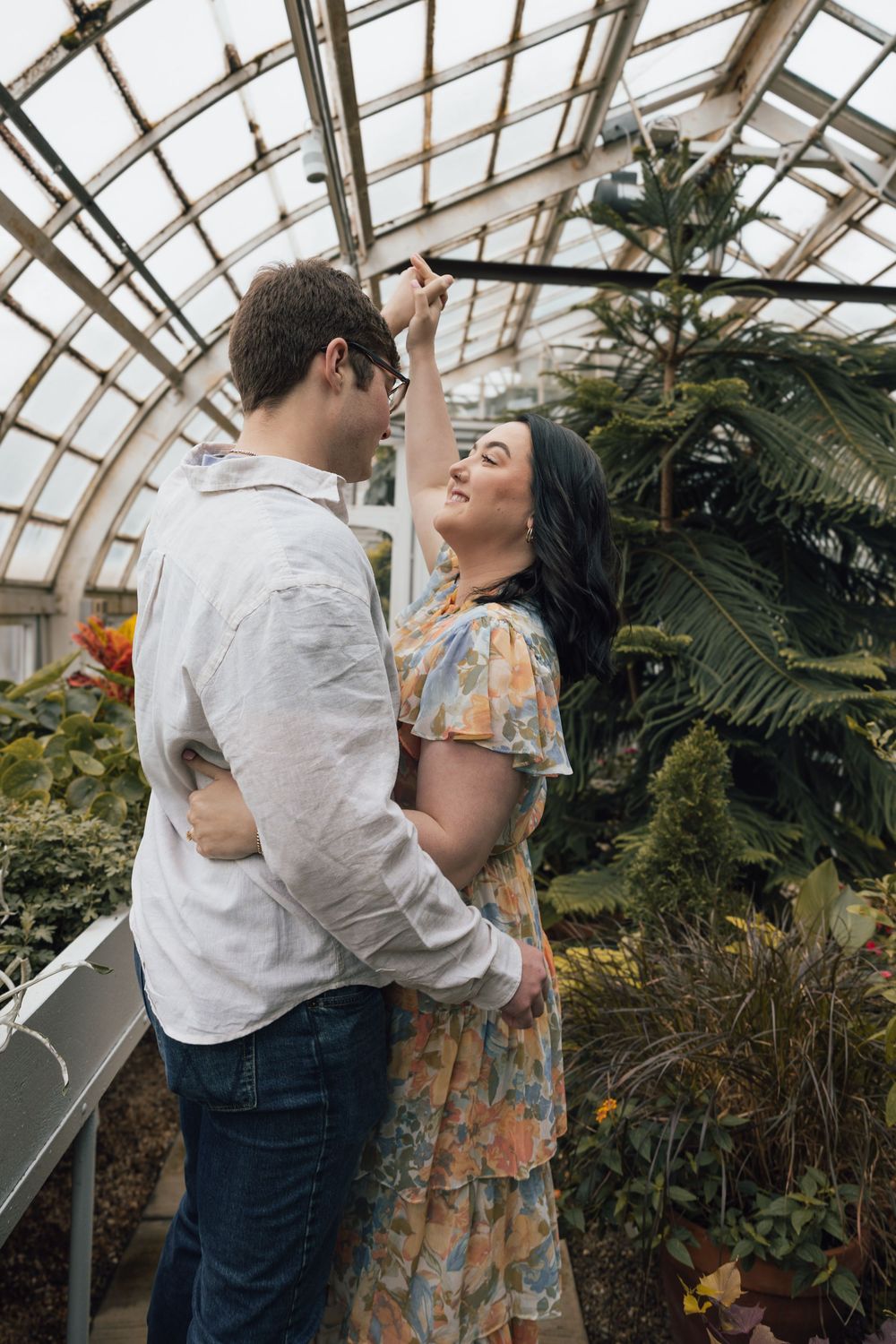 A romantic couple shares an intimate moment together inside a greenhouse filled with lush tropical plants and palm trees.