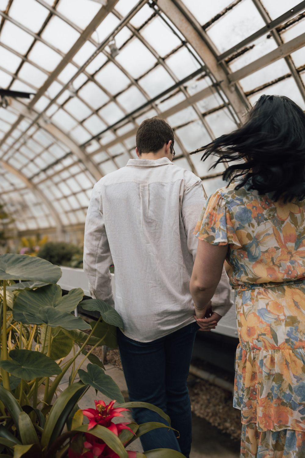 A couple explores a greenhouse together with a floral dress and white shirt visible among tropical plants and glass ceiling.