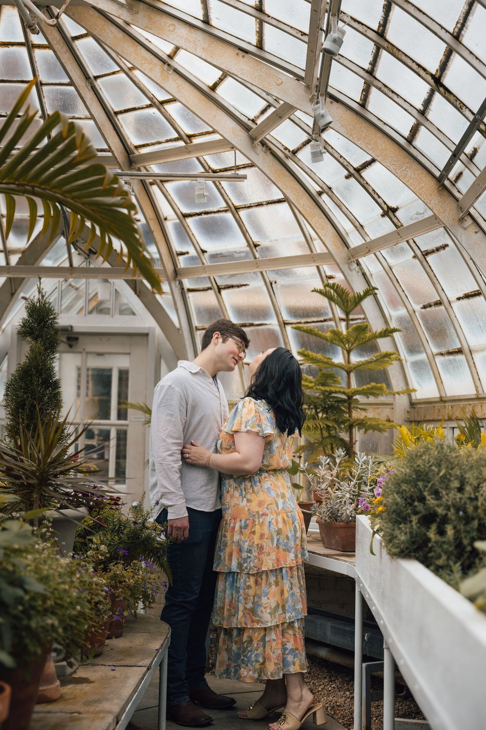 A romantic moment between a couple embracing in a sunlit greenhouse filled with tropical plants and greenery.