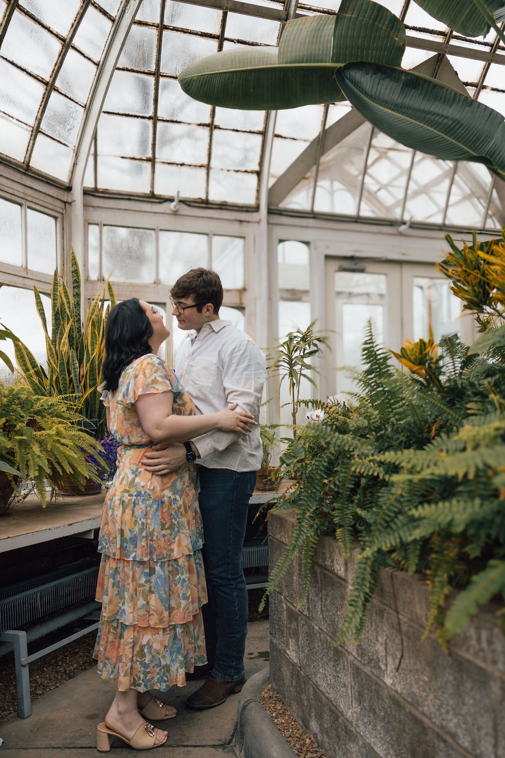 Romantic couple embracing in a sunlit greenhouse surrounded by lush ferns and tropical plants.