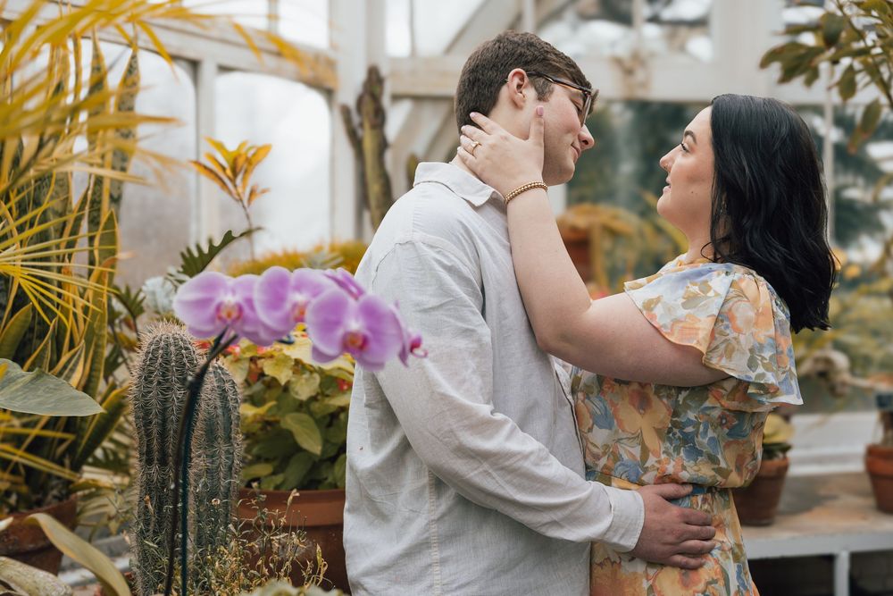 A romantic couple embraces in a greenhouse setting with blooming orchids and tropical plants surrounding them.