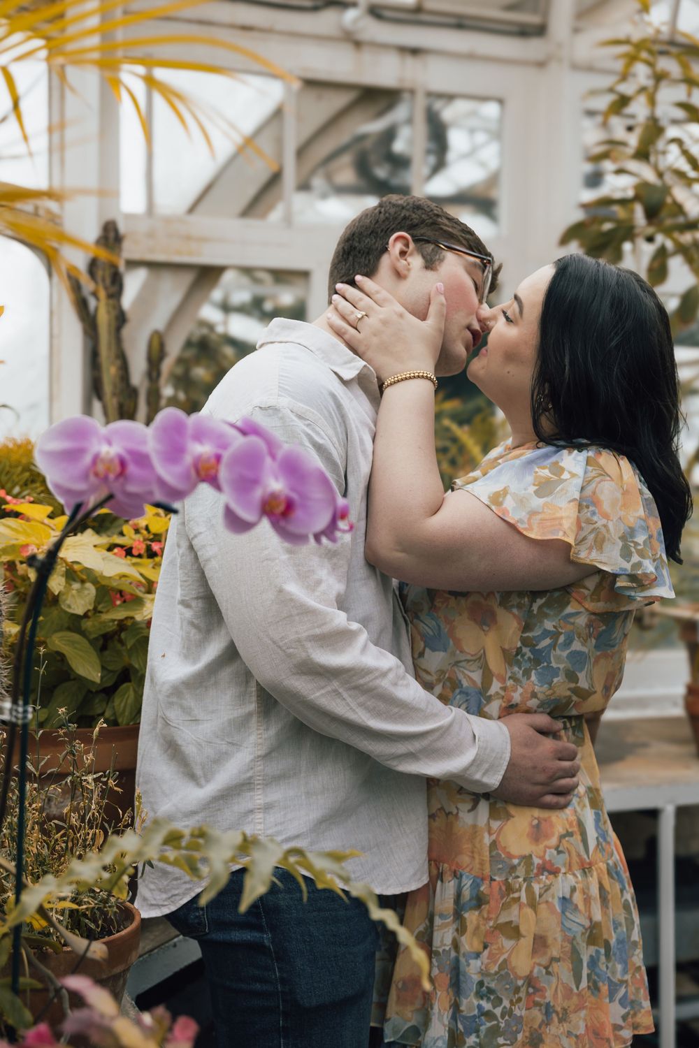 Romantic couple shares a tender kiss surrounded by orchids and tropical plants in a sunlit greenhouse.