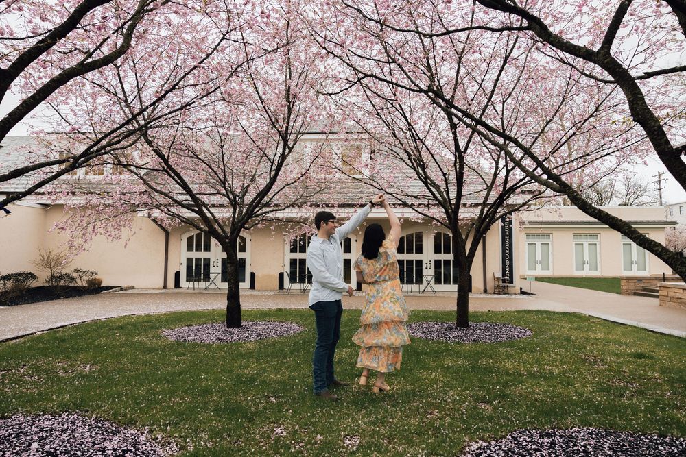 Two people play giant Jenga blocks under blooming cherry blossom trees outside a beige building.