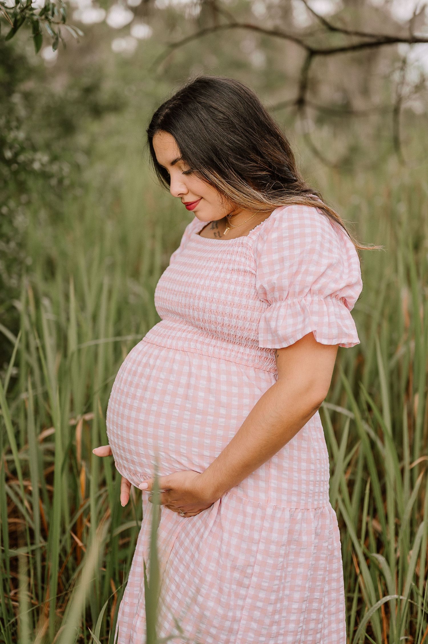 Expectant mother in pink gingham dress poses in tall grass during sunset.