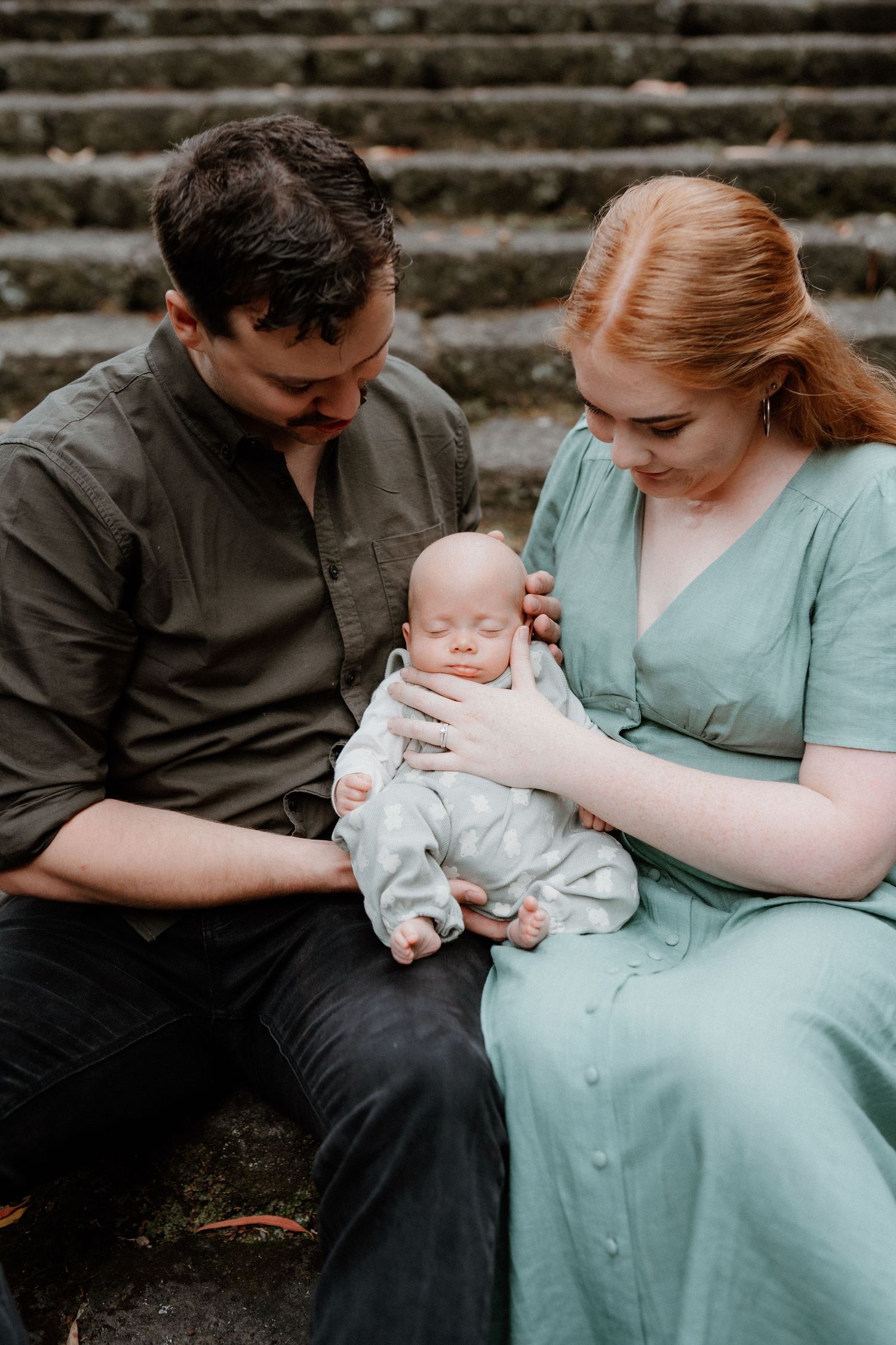 A couple in casual clothing sits on stone steps while tenderly holding their sleeping newborn baby.