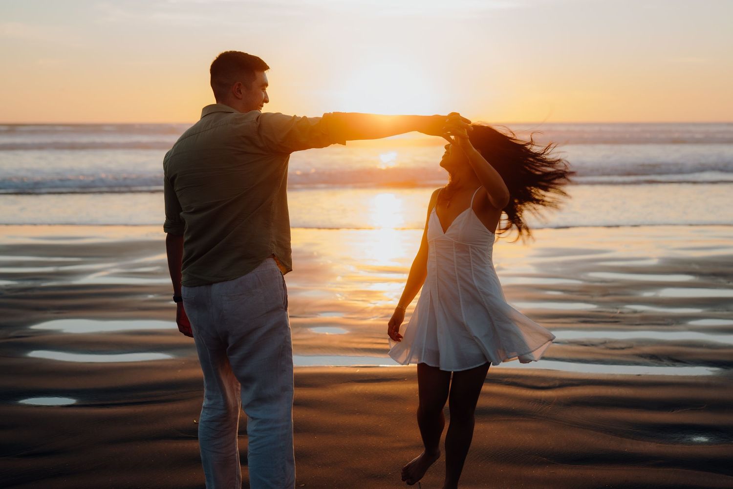 A silhouetted couple dances on the beach at sunset with ocean waves in the background.