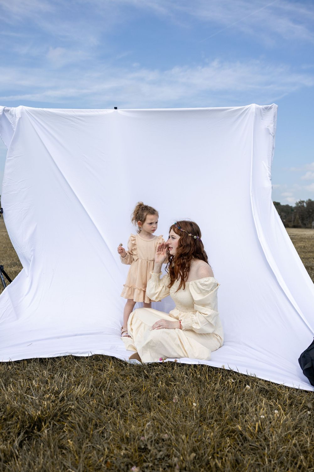 A sweet outdoor photo session with a white backdrop featuring two people in cream-colored dresses sitting on grass.