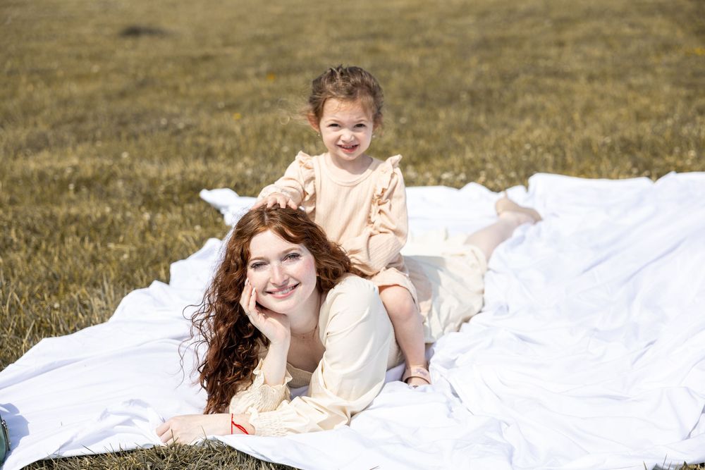 Two people in cream-colored outfits lying on a white blanket in a grassy field on a sunny day.