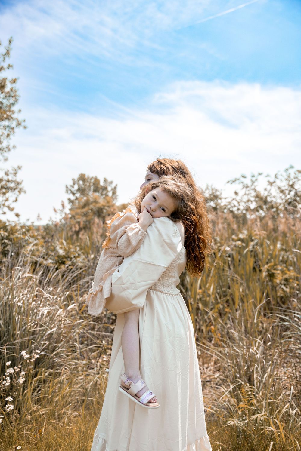 Mother and daughter share a tender embrace in a golden field of tall grasses under a bright blue sky.