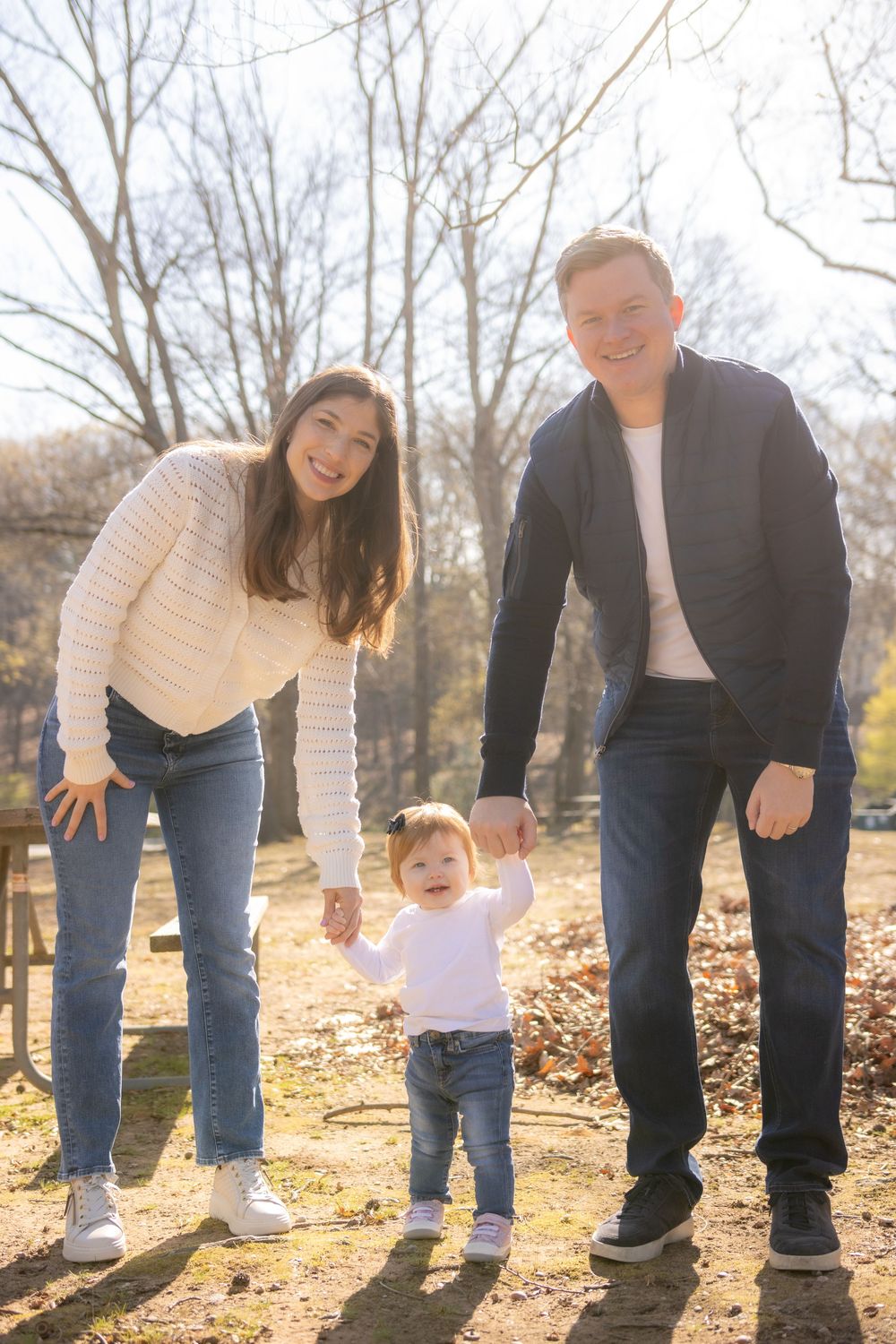 Family enjoying a sunny autumn day outdoors with their toddler walking between parents on fallen leaves.
