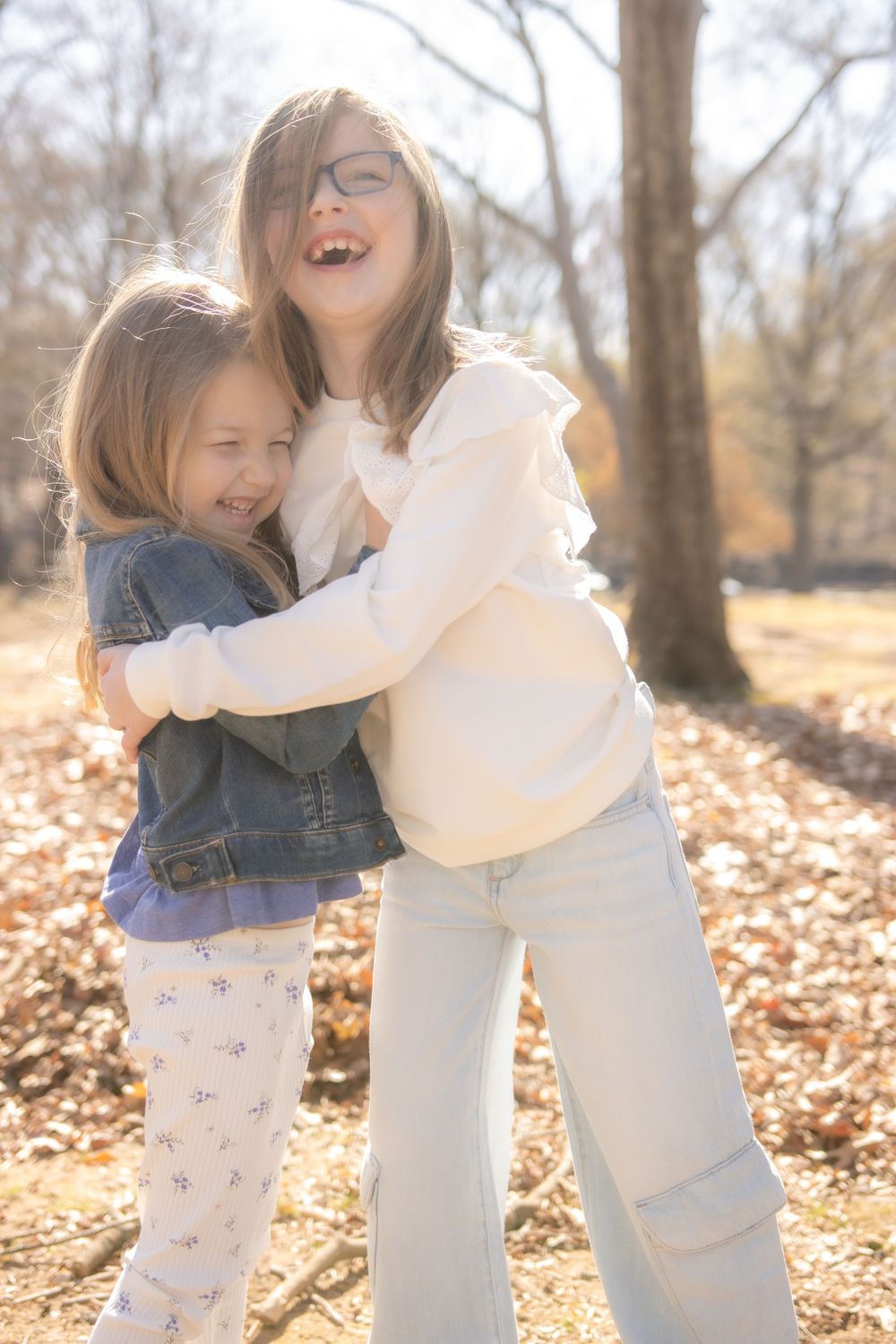 Two people share joyful laughter while embracing among fallen autumn leaves in a sunlit forest setting.