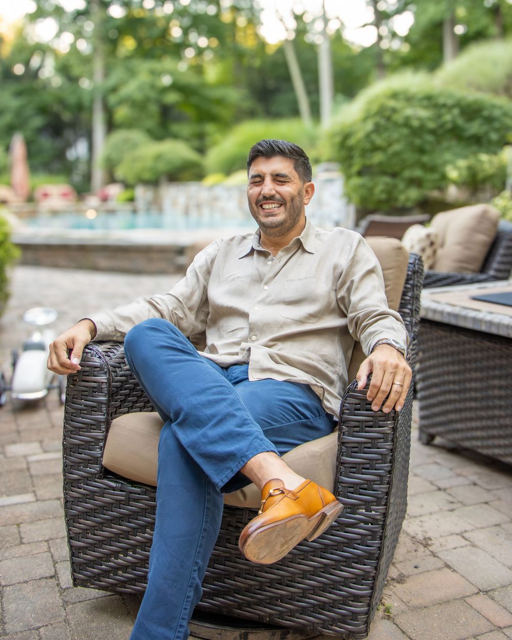 Person relaxing in a wicker patio chair wearing blue jeans and brown shoes on an outdoor terrace with garden furniture.