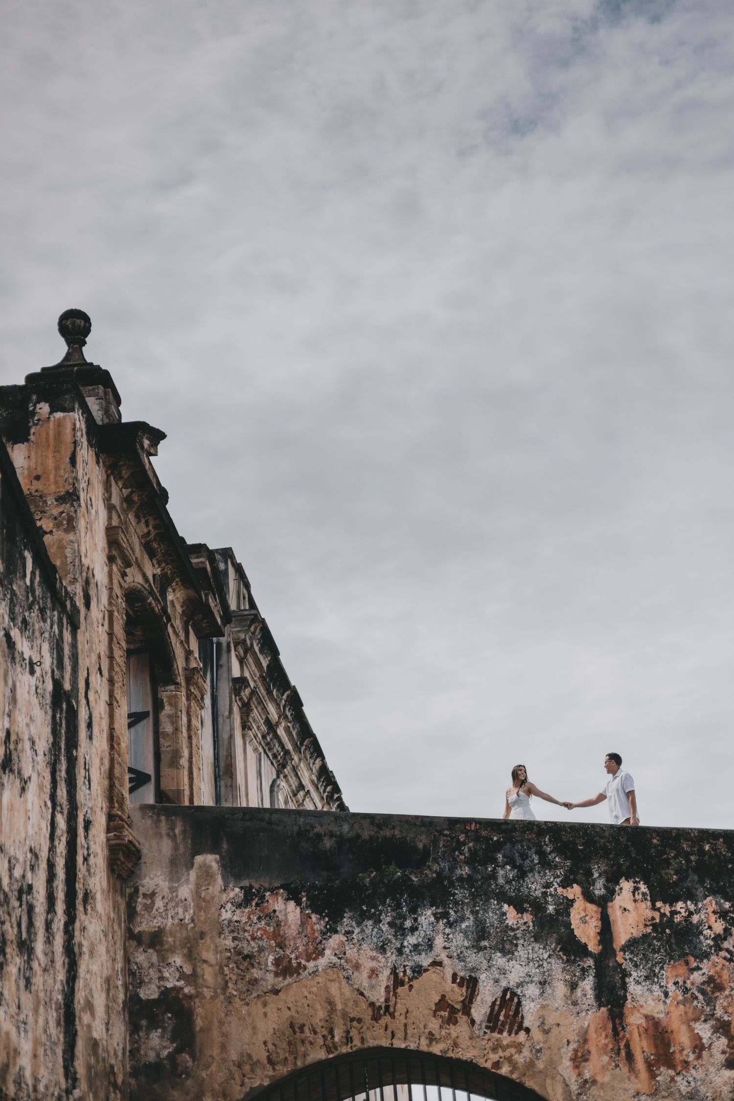 A couple in coastal wedding attire stands holding hands by the ocean while gazing in opposite directions.