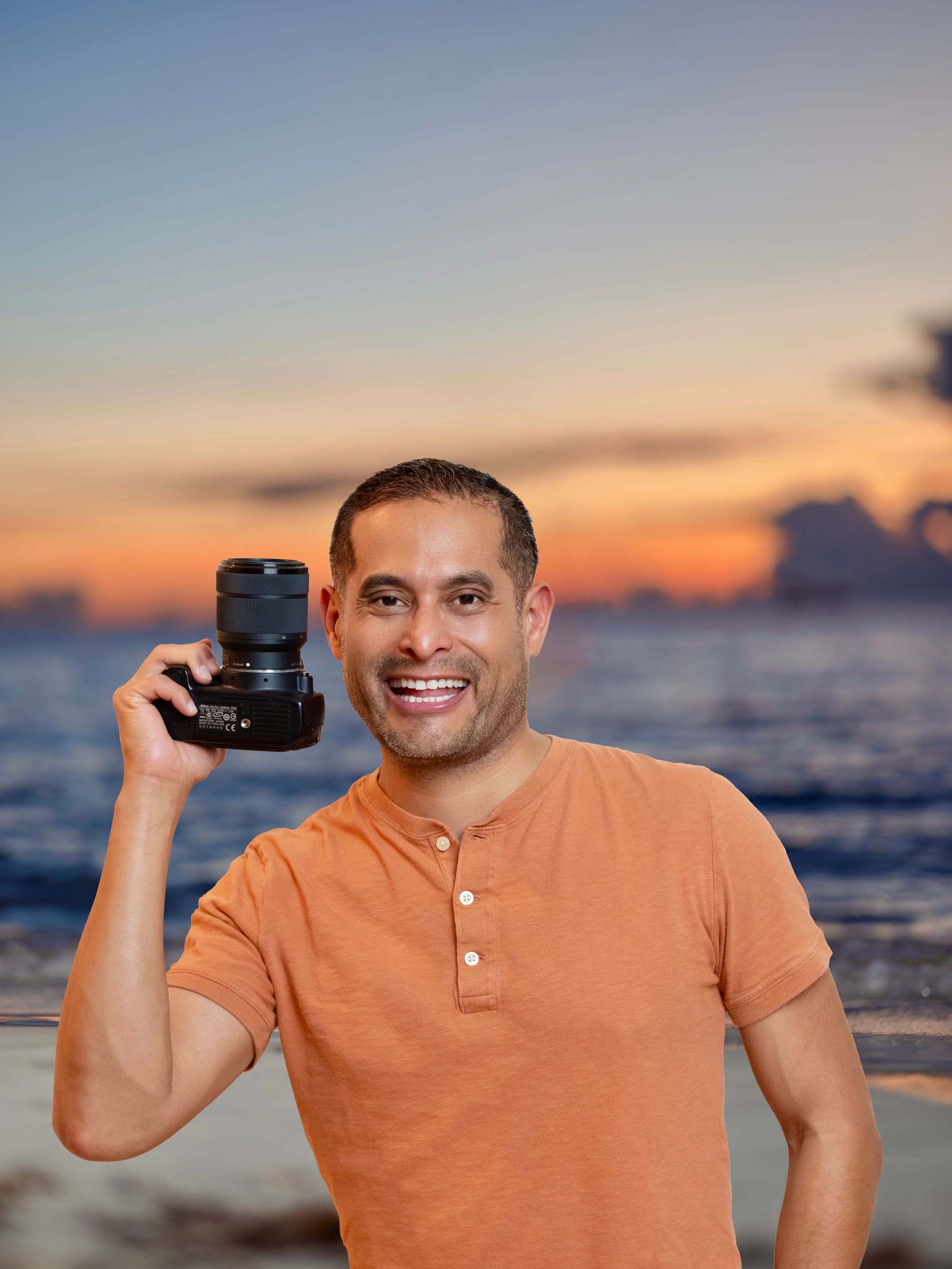 John Burgos - YonnBurgos in orange shirt holding a camera with an ocean sunset background