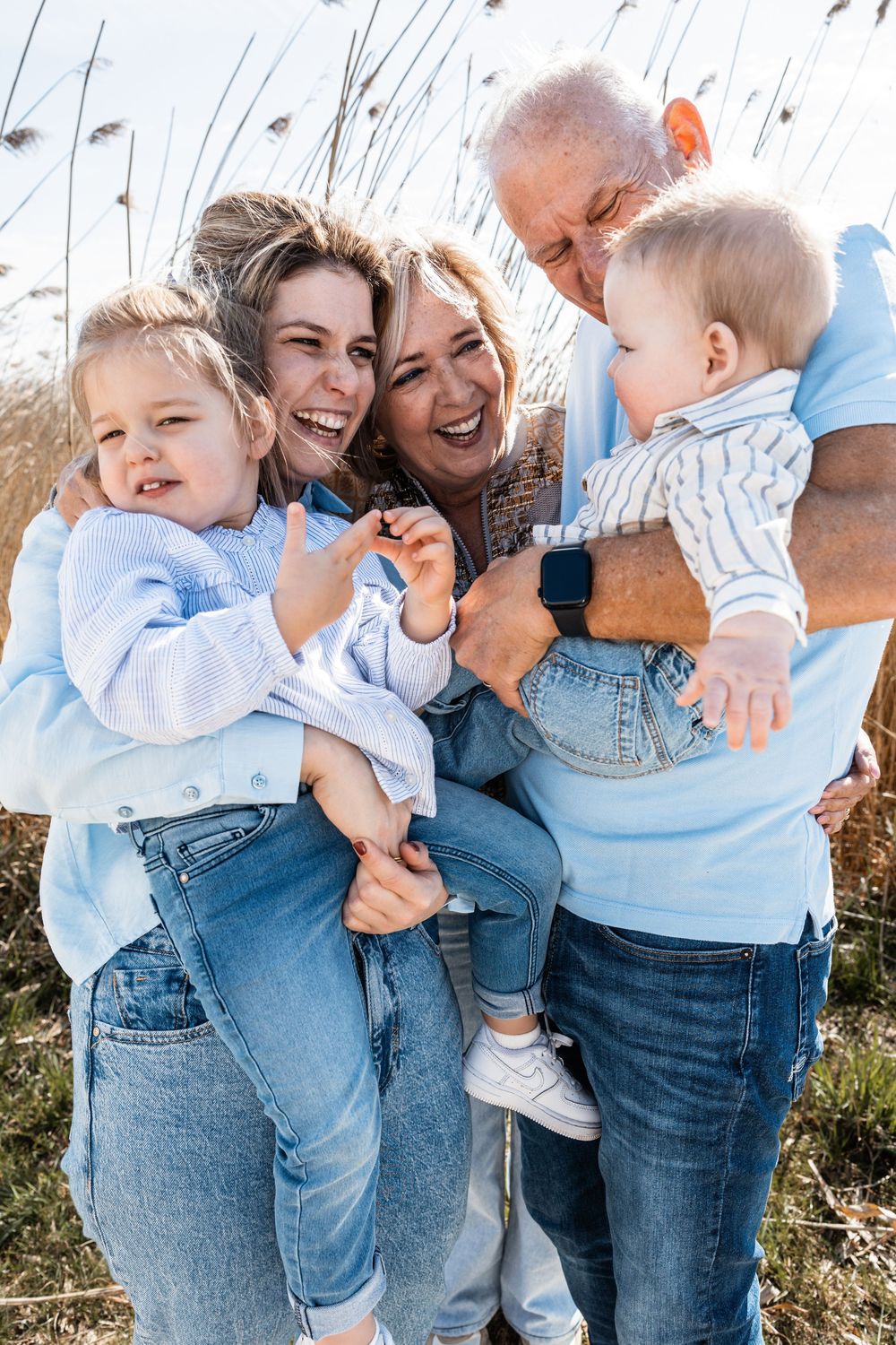 Happy family wearing matching denim and blue outfits laugh together outdoors among tall grass on sunny day.