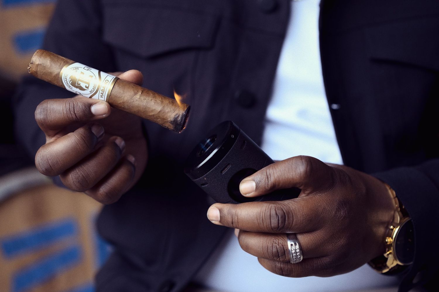 Hands holding a lit cigar and mobile phone while wearing a black suit and gold jewelry.