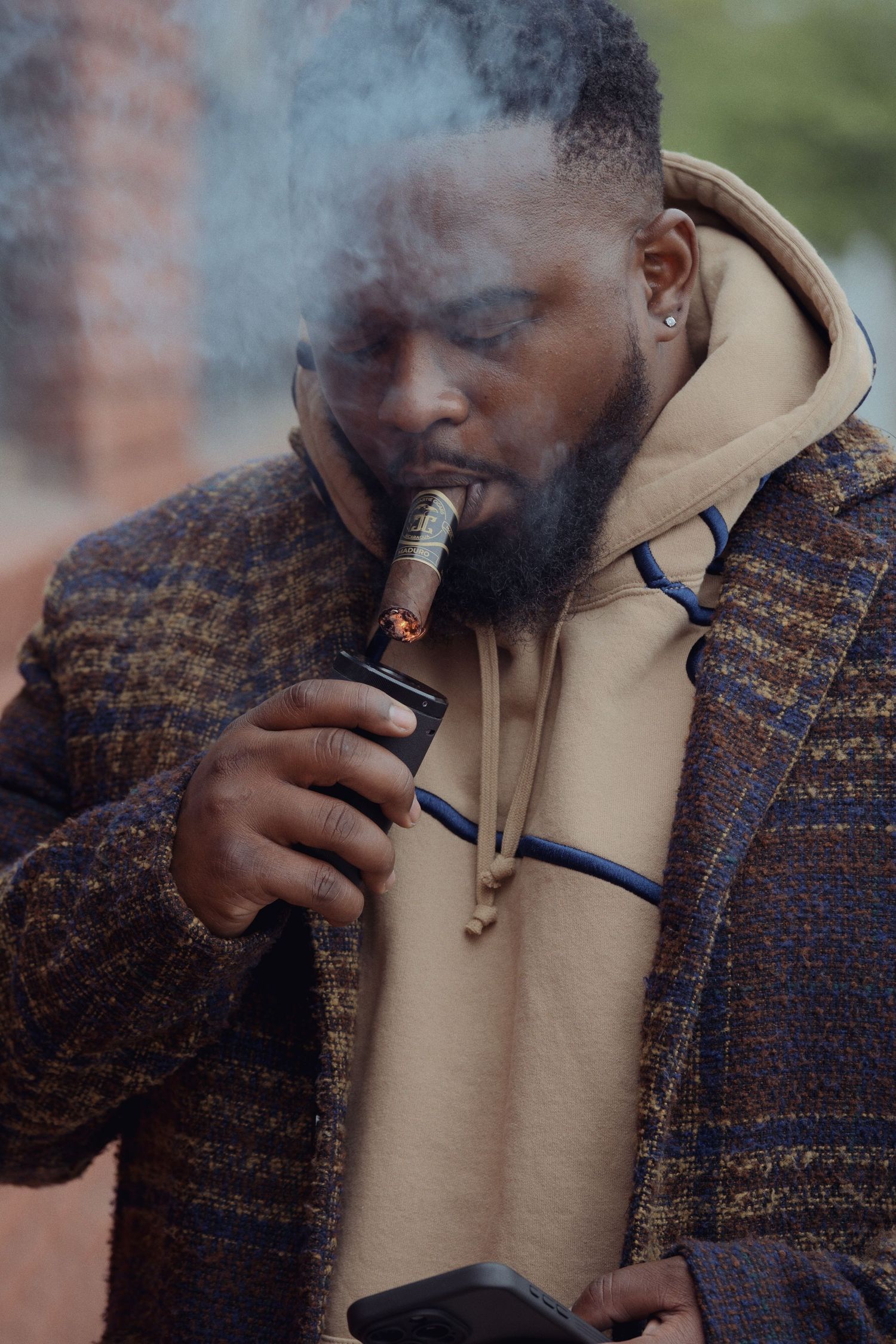 Person wearing a brown plaid coat and beige hoodie lights a cigar while smoke drifts in the air.