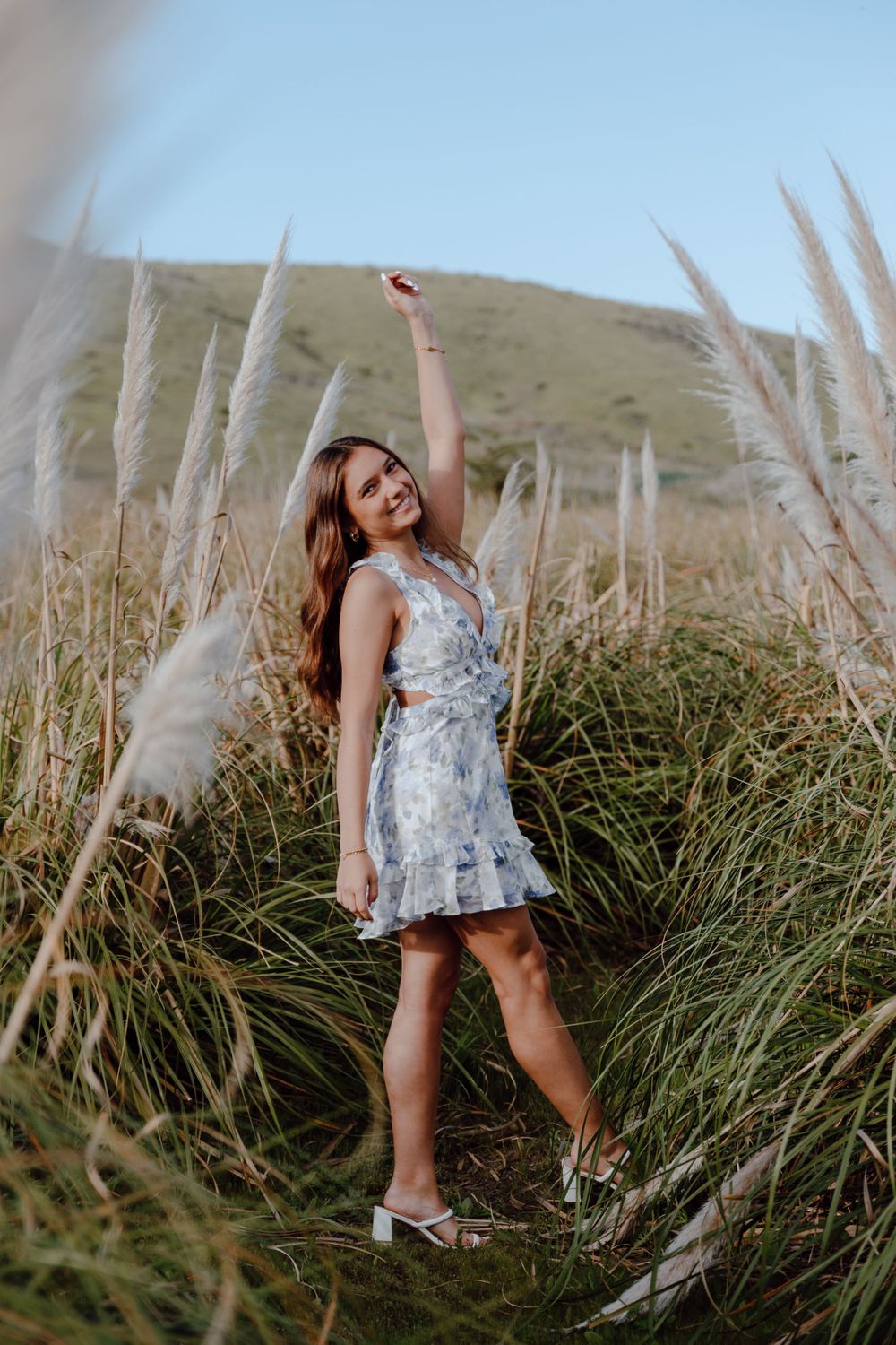 Person in floral dress twirling joyfully among tall grass with mountains in background.