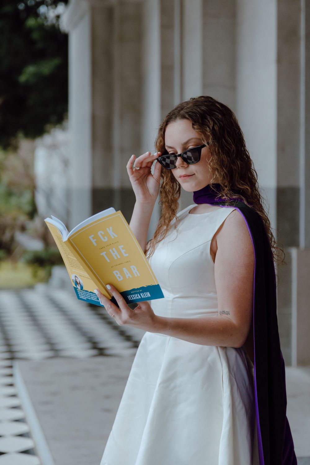 A person in a white dress and purple cape reads a yellow book on a columned porch.