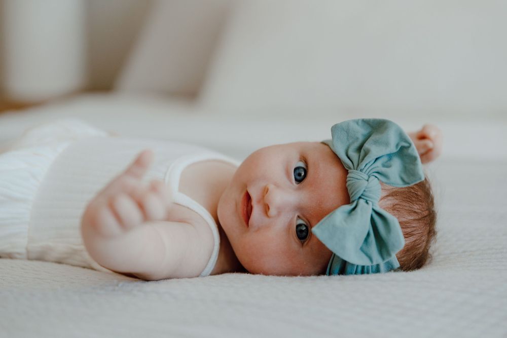 Baby in white outfit with teal bow headband lying on soft white bedding.