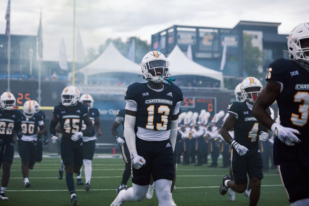 A football player in dark uniform stands on the field during pre-game preparations.