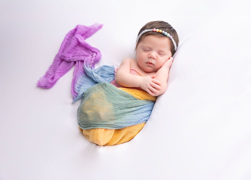 Sleeping newborn wrapped in colorful fabrics against white background.
