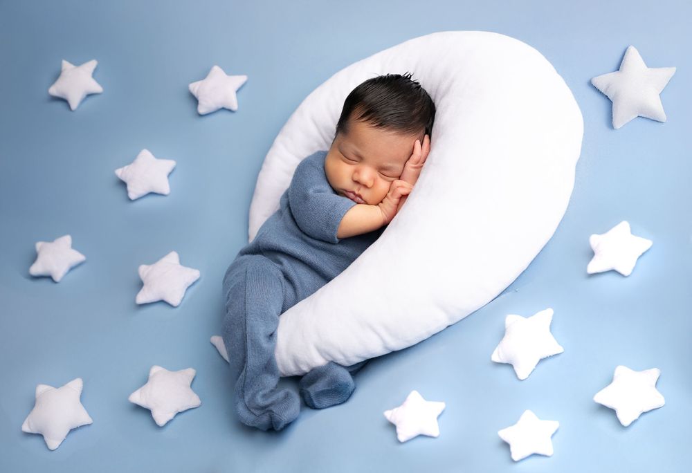 Sleeping newborn in blue wrap on crescent moon pillow with decorative stars.