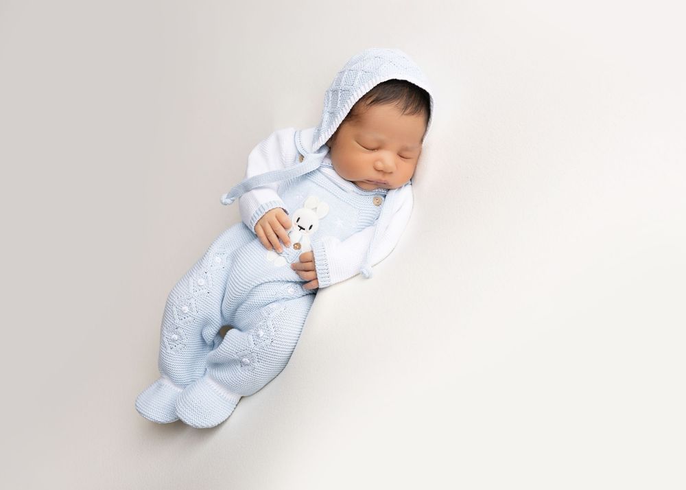Sweet newborn baby dressed in light blue sleeper rests peacefully on white blanket.