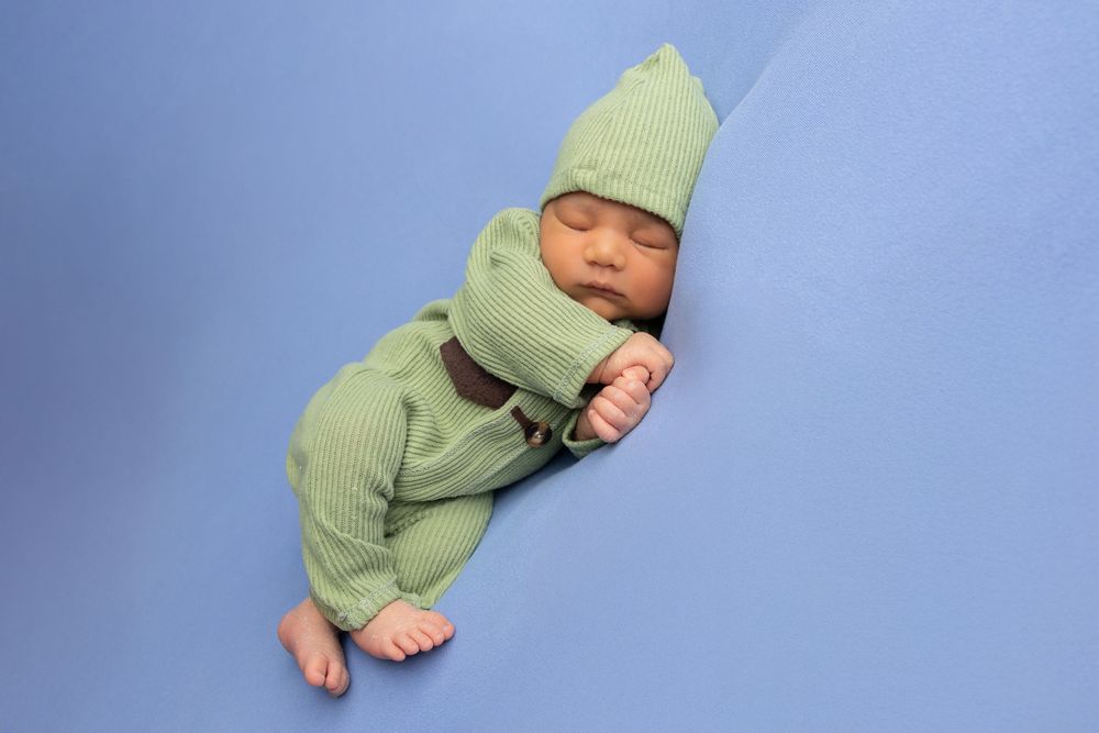 Sleeping newborn baby wearing soft mint green outfit and hat rests peacefully on blue background.