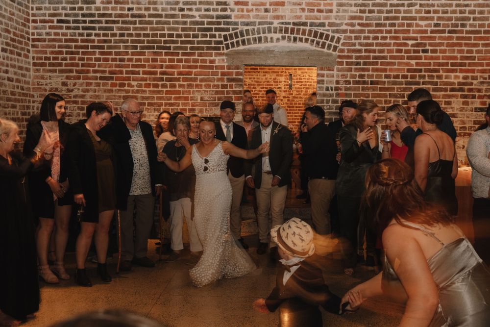 Wedding reception celebration in a rustic brick-walled venue with guests gathered around the dance floor.