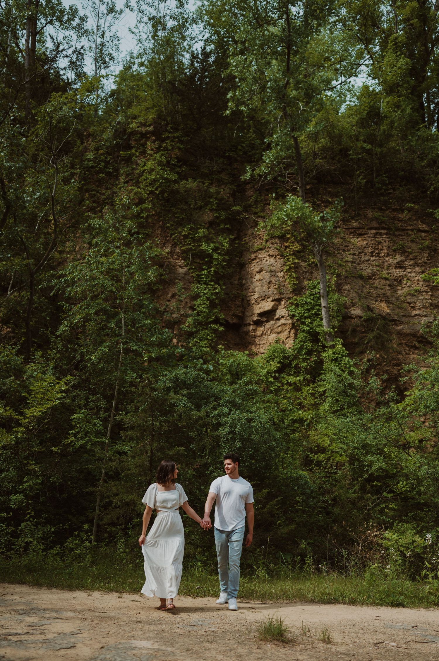 Two people in white and blue outfits hold hands while walking along a forested trail.