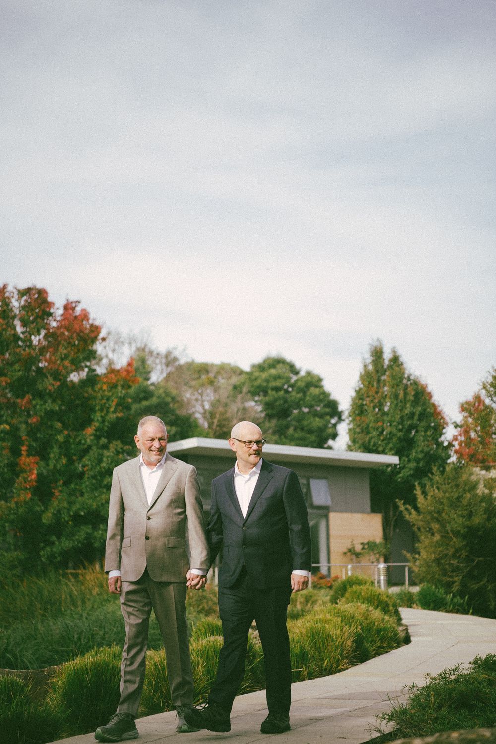 Two people in suits walking hand in hand on a garden path surrounded by autumn foliage.