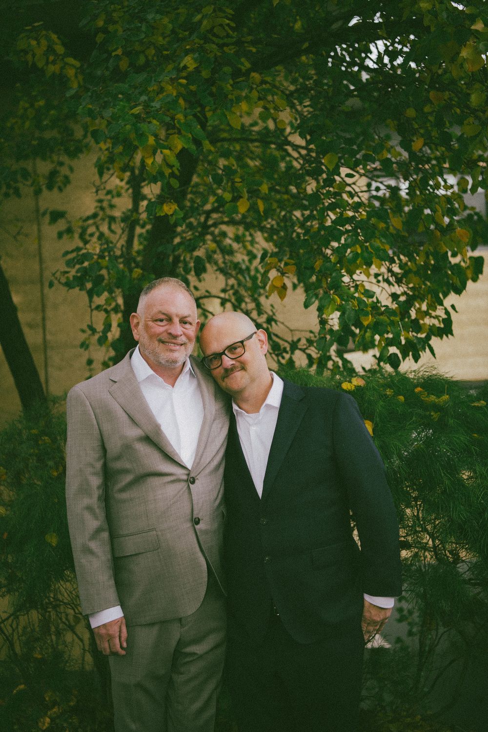 Two people in contrasting suits pose together under tree branches in a garden setting.