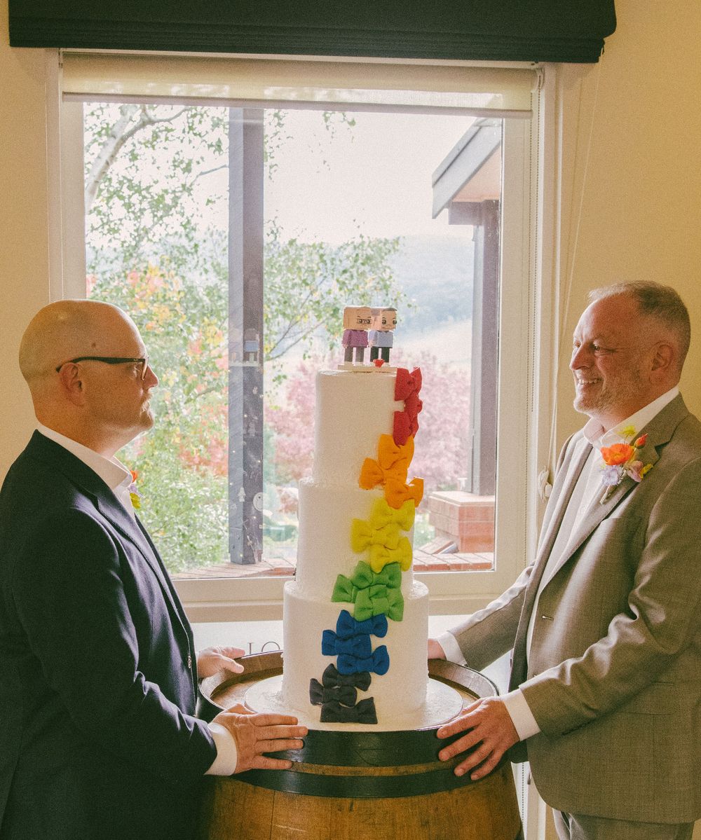 Two people stand beside a tall white wedding cake decorated with rainbow-colored puzzle pieces.