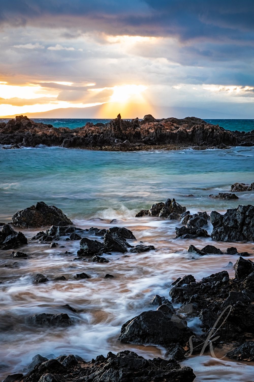 Landscape photo of sun bursting through the clouds on the rocks as the waves break in Kihae, Maui Hawii.