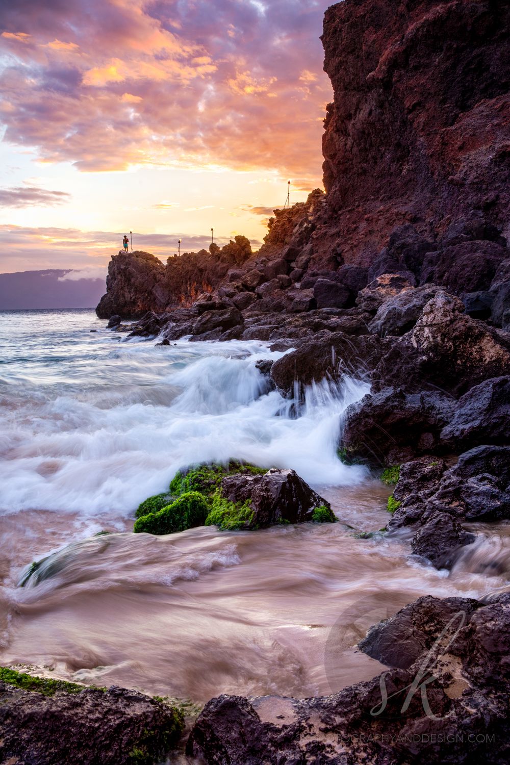 Landscape photo of waves crashing along the black rocks of Kaanapali Beach, Maui and evening torches lit.