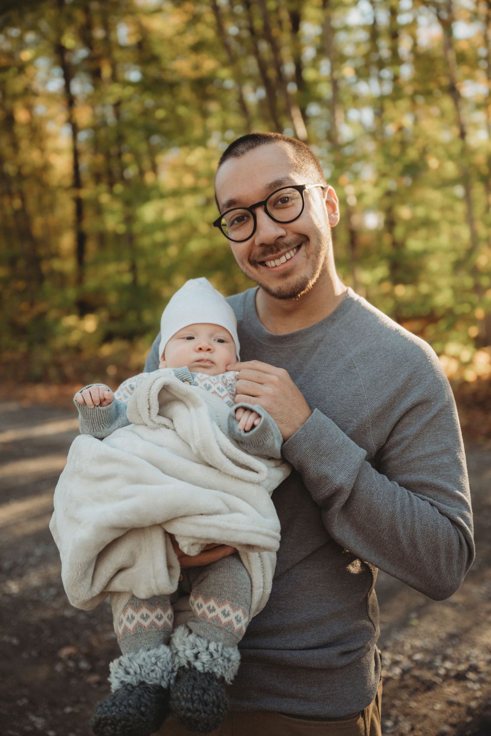 A parent in a grey sweater holds a bundled baby wearing a white hat in a forest setting.