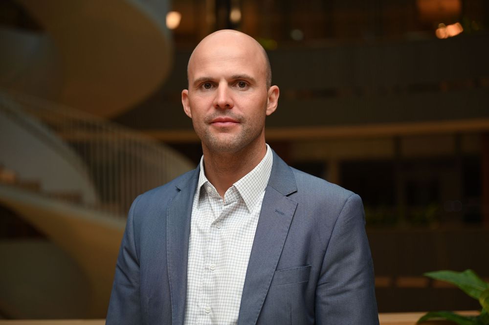 Professional headshot of person wearing a grey suit jacket and white dress shirt in an office setting with warm lighting.