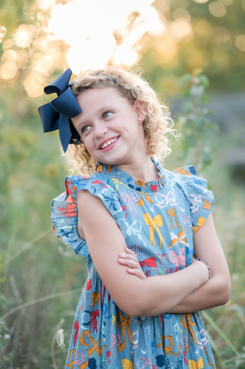 Young child in blue floral dress with ruffled sleeves and navy hair bow smiling in outdoor garden setting at sunset.