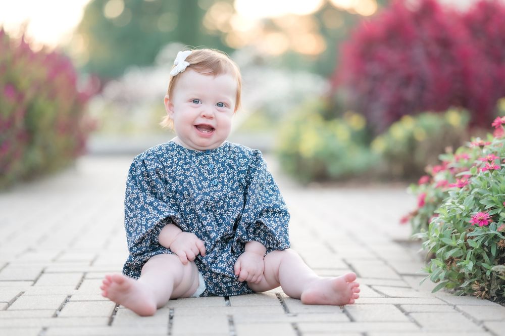 A smiling baby wearing a blue floral dress sits on a stone path surrounded by colorful garden flowers.