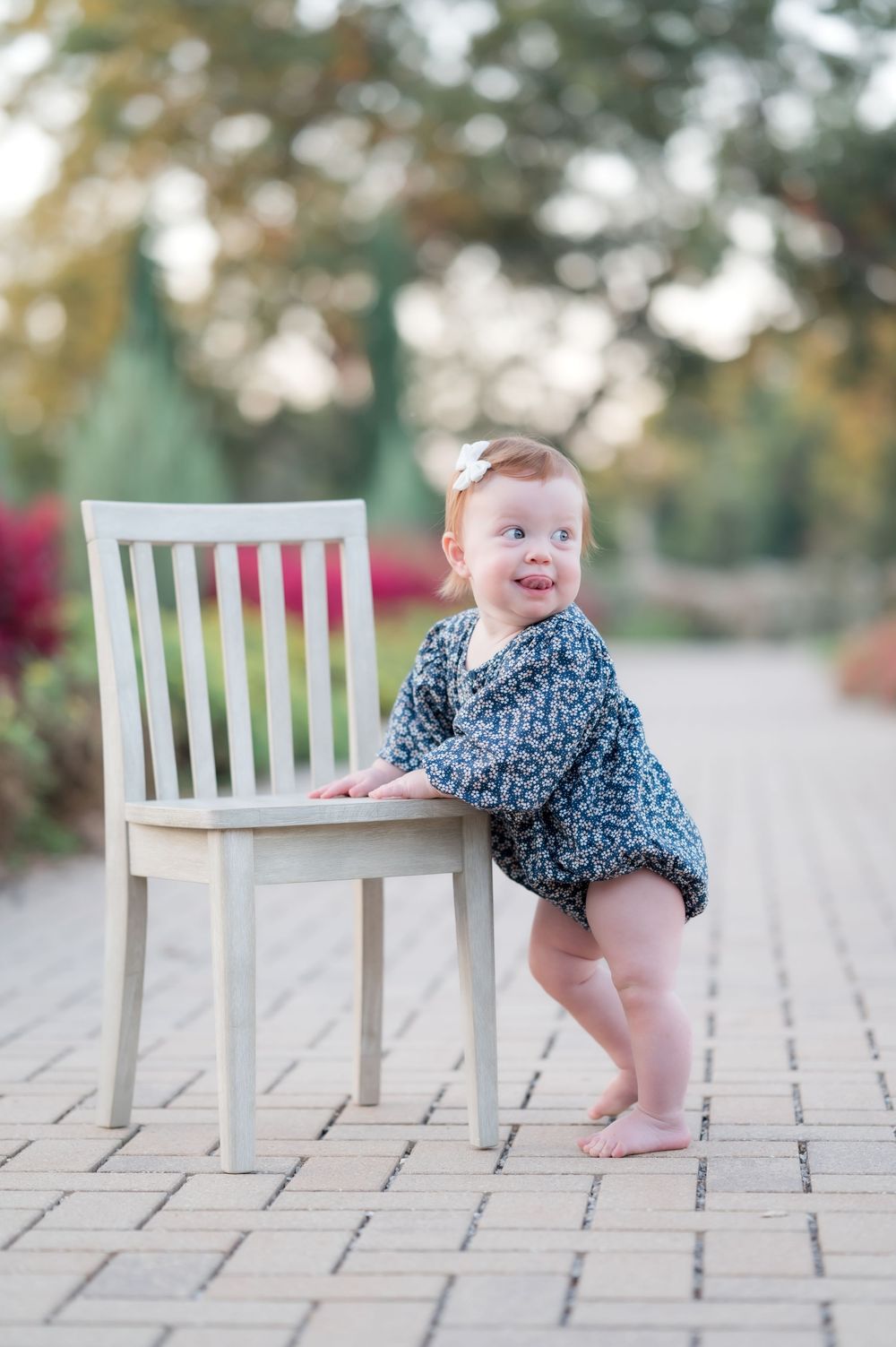 A small child in a blue patterned top stands smiling next to a white wooden chair on a tiled outdoor patio.