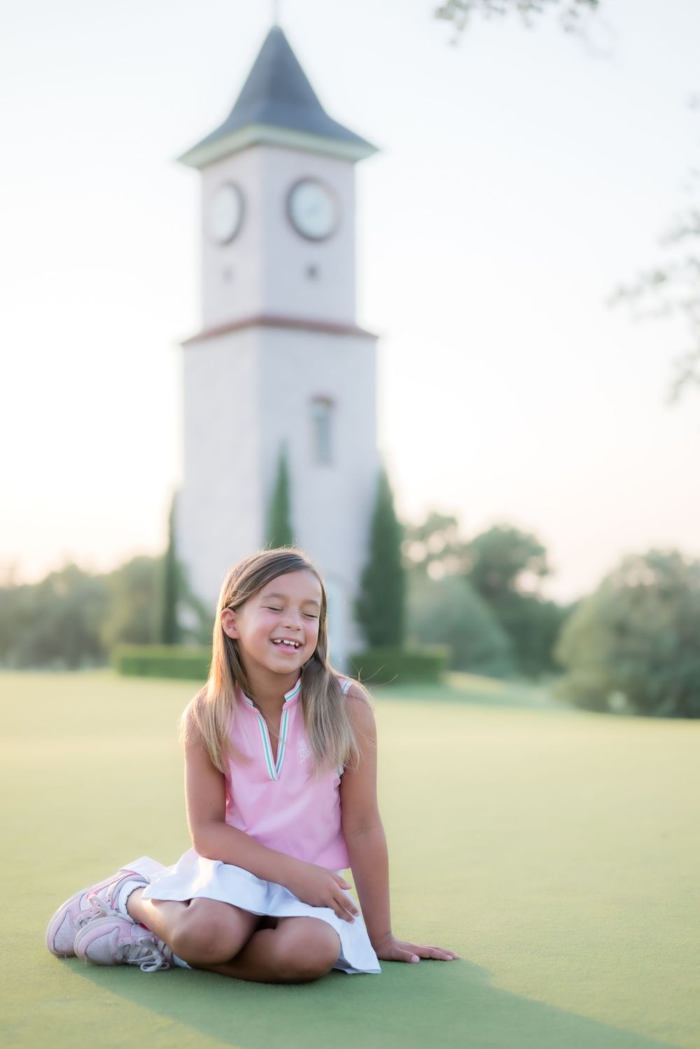 A golf enthusiast sits on a putting green at sunset with a white clock tower visible in the background.
