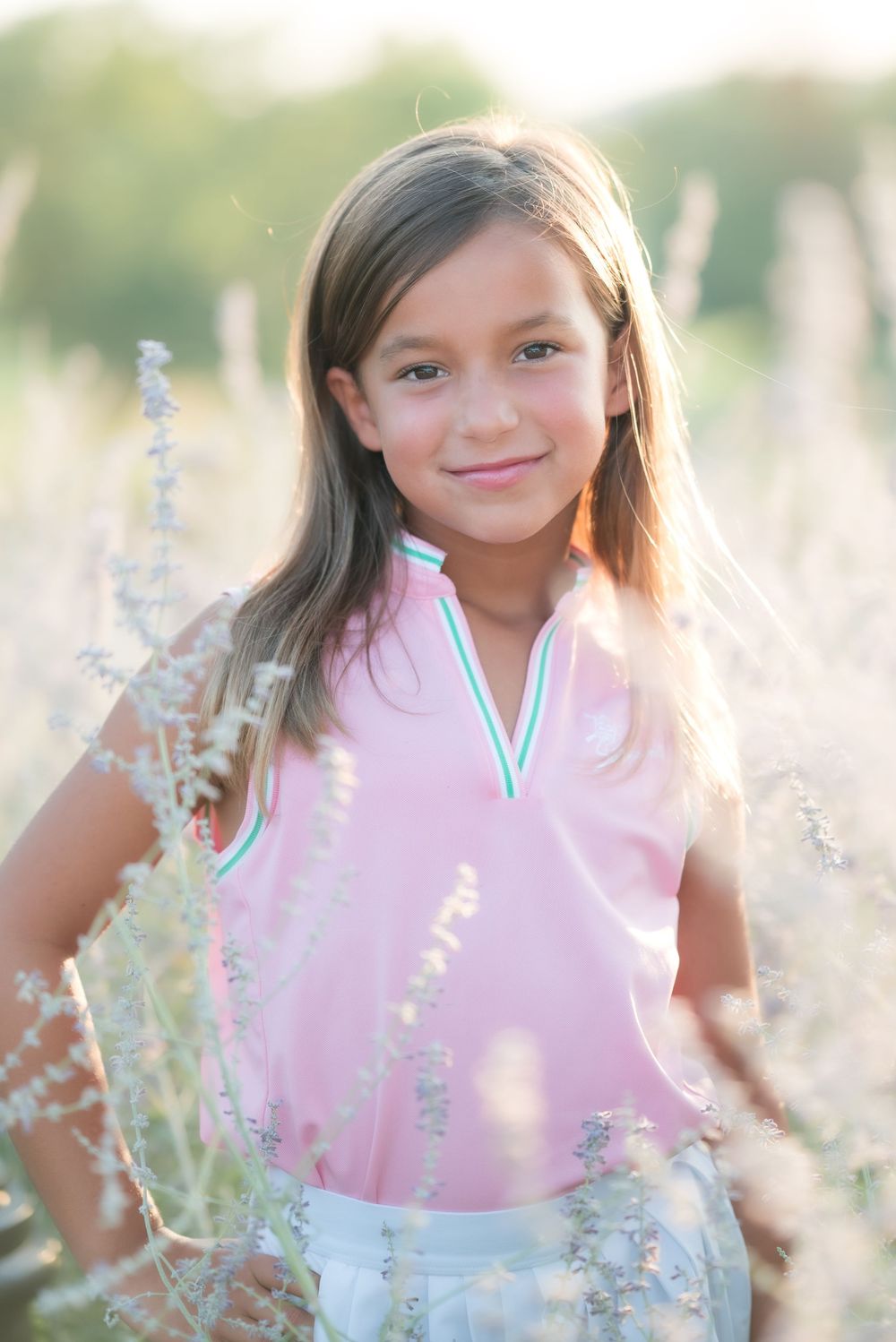 A child in a pink v-neck top poses outdoors among delicate wildflowers at golden hour.