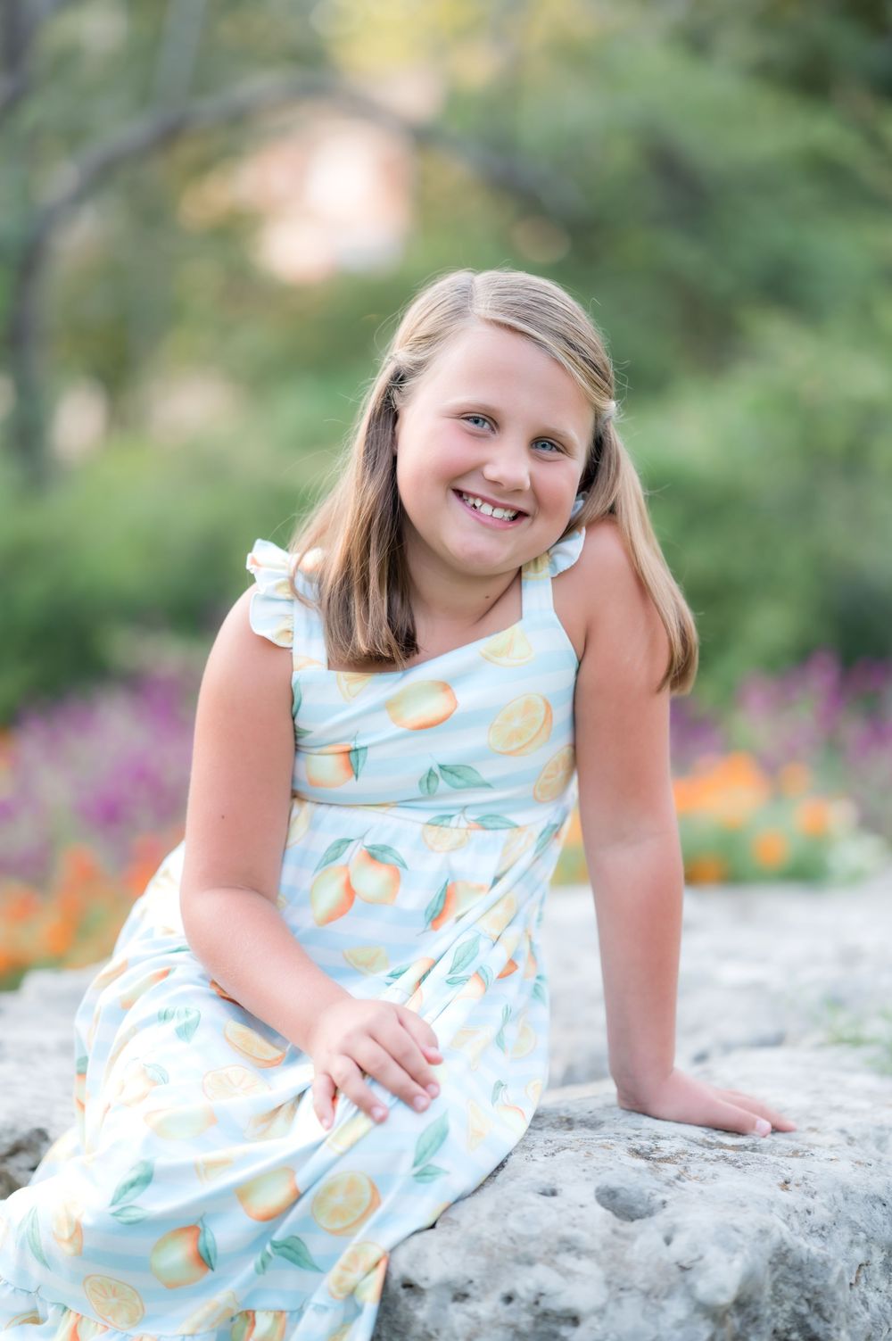 Young girl in a light blue patterned dress sits on a rock with colorful flowers blooming in the garden background.