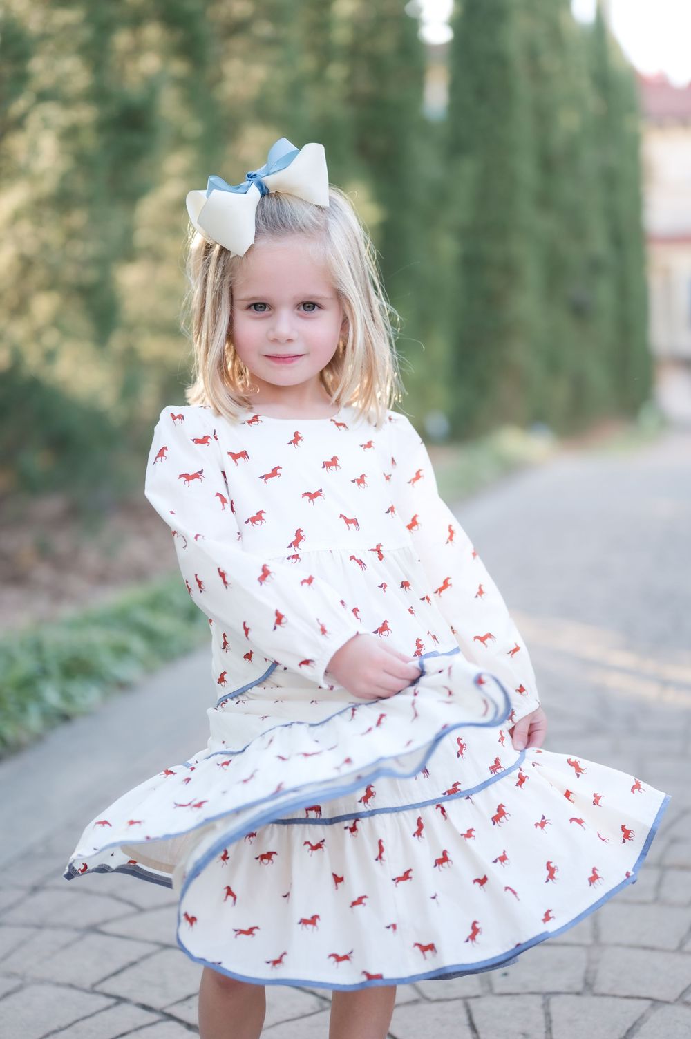 White dress with red bow pattern and blue trim twirls on a stone path with cypress trees in background.