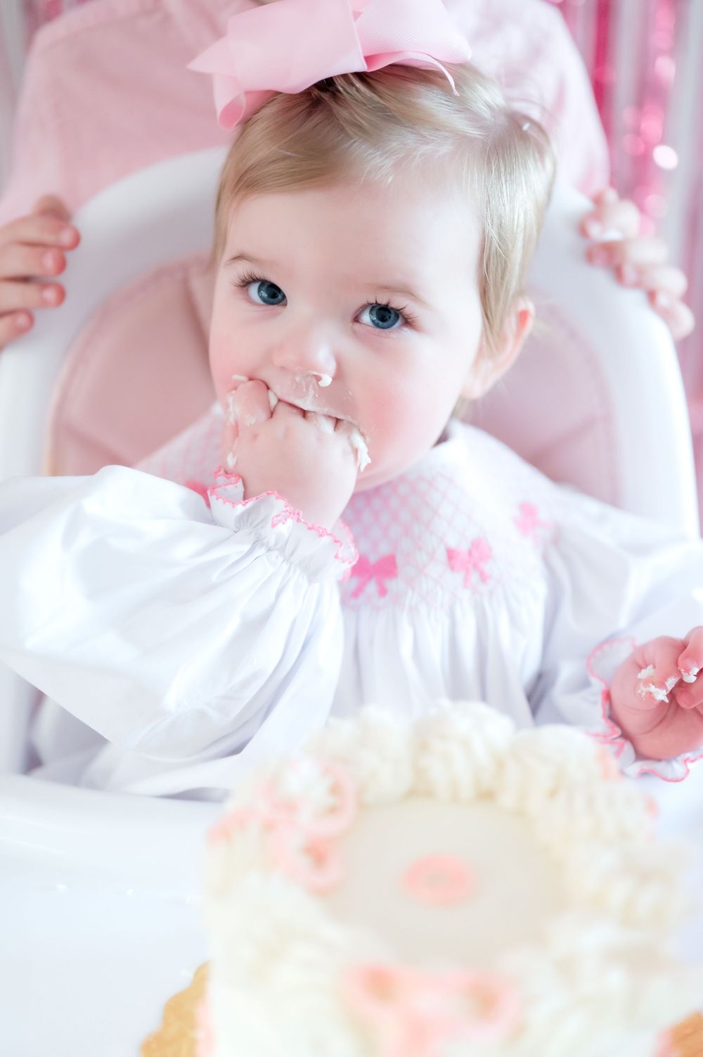 Baby in white smocked dress with pink bows sits in high chair with pink decorations and party decor.